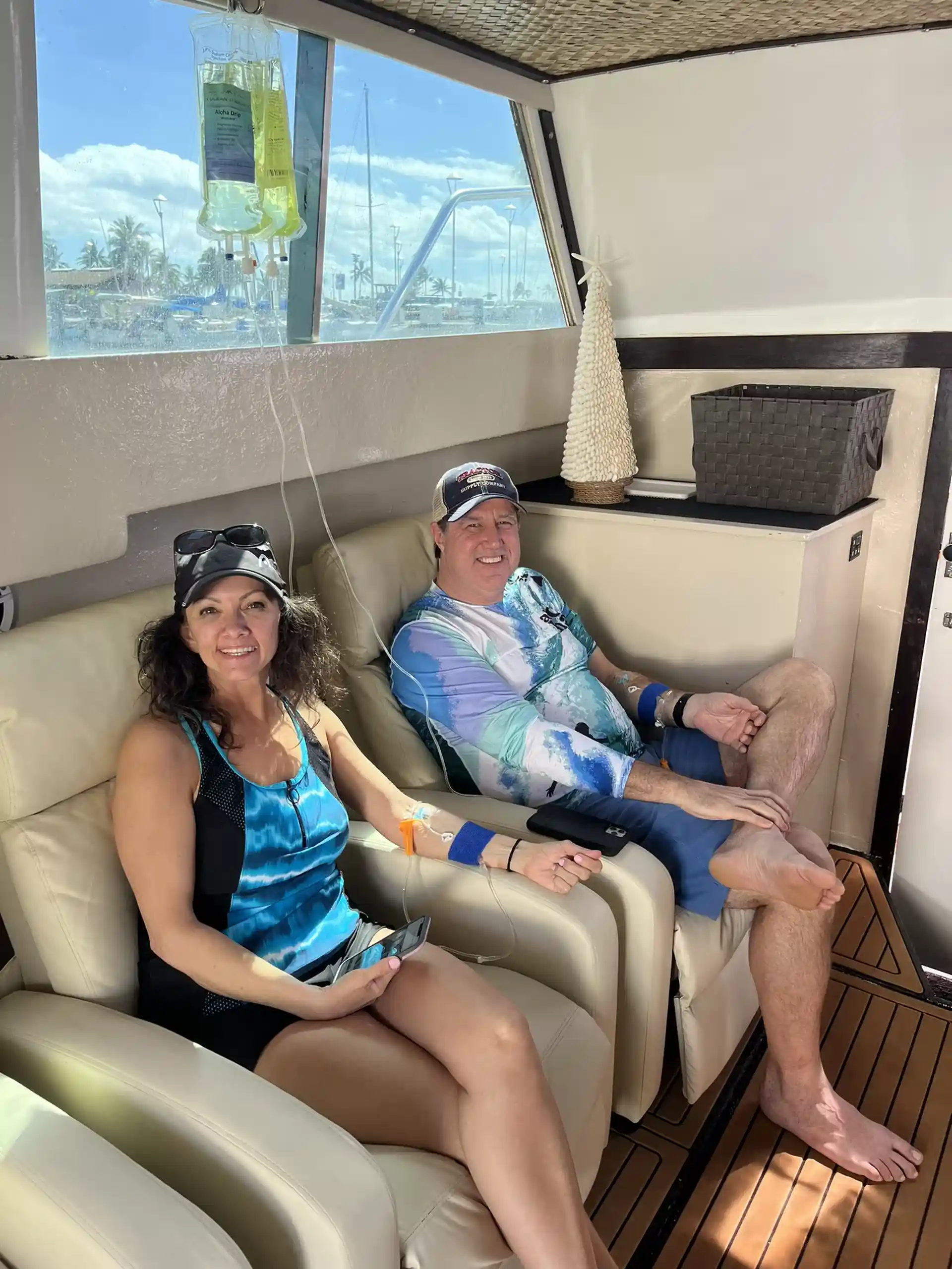 The photo shows two people sitting comfortably in a boat cabin, receiving IV therapy. Both are smiling and appear relaxed. The woman is wearing a blue top and black shorts, while the man wears a cap and a patterned shirt. IV bags hang next to them